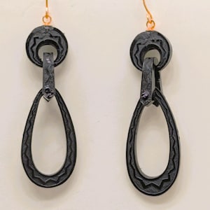 May include: A pair of black drop earrings with a textured design. The earrings have a circular top with a decorative pattern, connected to a teardrop-shaped dangle. The ear wires are gold-colored.