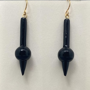 Peut inclure: Une paire de boucles d'oreilles en bois noir avec une boule ronde et une pointe pointue. Les boucles d'oreilles sont suspendues à des crochets dorés.