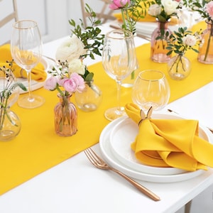 Puede incluir: Un camino de mesa amarillo con flores blancas en pequeños jarrones de vidrio. La mesa está puesta con platos blancos, cubiertos dorados y servilletas amarillas con anillos de servilleta de madera.