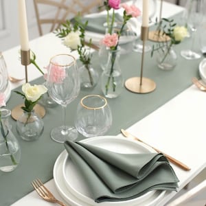 Puede incluir: Una mesa puesta con un camino de mesa verde salvia, platos blancos y cubiertos dorados. La pieza central presenta pequeños jarrones con flores rosas y blancas y candelabros altos con velas blancas.