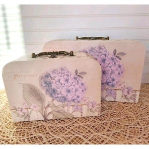 May include: Two vintage-style, decorative wooden boxes with floral designs. The boxes are painted in a light cream color and feature a floral pattern with purple hydrangeas. The boxes have a hinged lid and a brass handle.