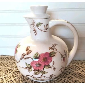 May include: A white ceramic pitcher with a floral design featuring pink roses and brown leaves. The pitcher has a lid and a handle.