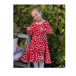 Könnte beinhalten: Ein junges Mädchen trägt ein rot-weiß gepunktetes Kleid mit langen Ärmeln. Das Kleid hat einen Rüschen-Saum und einen weißen Besatz.