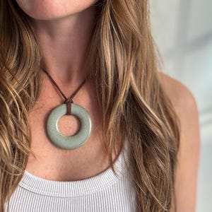 May include: A light green ceramic pendant necklace with a brown leather cord. The pendant is a large, round ring with a hole in the center.