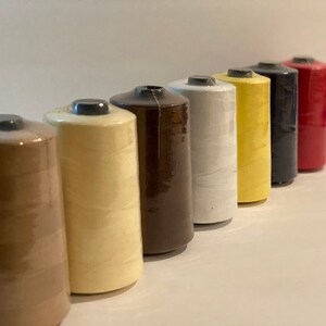 May include: Seven spools of thread in various colors, including brown, beige, white, yellow, black, and red. The spools are arranged in a row from left to right, with the brown spool at the left and the red spool at the right.