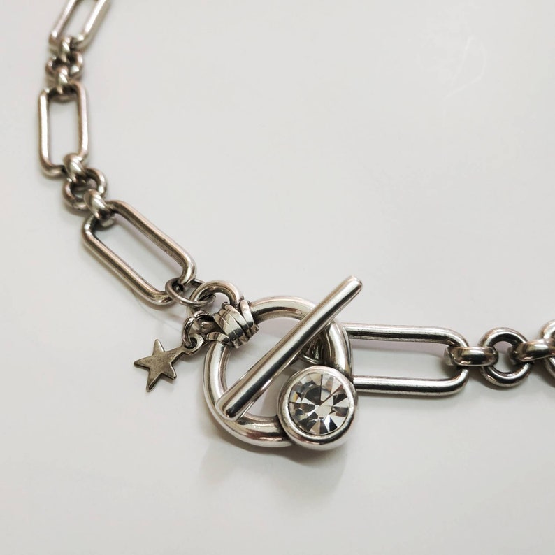 May include: A silver chain necklace with a toggle clasp featuring a clear rhinestone. The necklace has a star charm attached to the clasp.