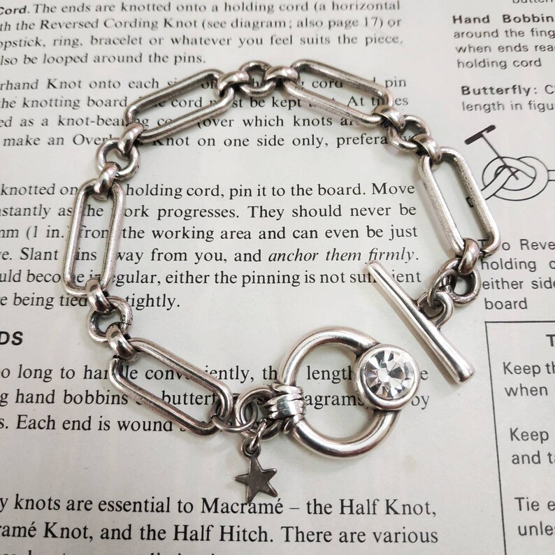 May include: A silver chain bracelet with a silver toggle clasp and a star charm. The bracelet has a clear crystal charm and is on a white background with text about macrame knots.