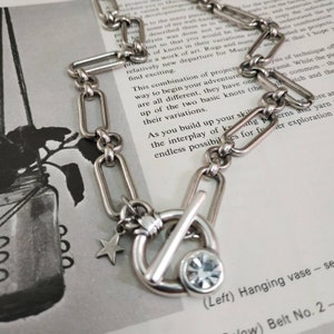 May include: A silver chain necklace with a toggle clasp, a star charm, and a clear rhinestone charm. The chain is made of rectangular links.