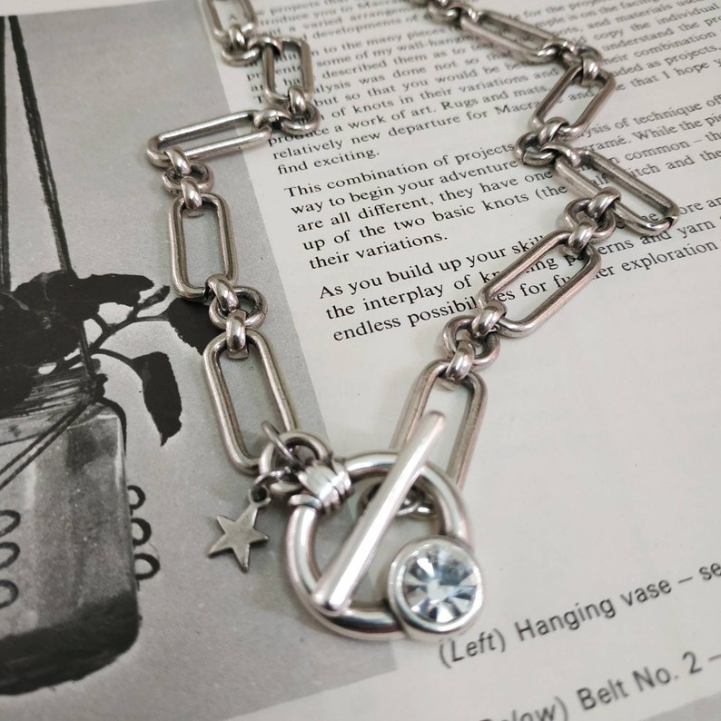 May include: A silver chain necklace with a toggle clasp, a star charm, and a clear rhinestone charm. The chain is made of rectangular links.