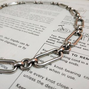 May include: A silver chain necklace with oval links. The chain is lying on a page of a book with text about knots.