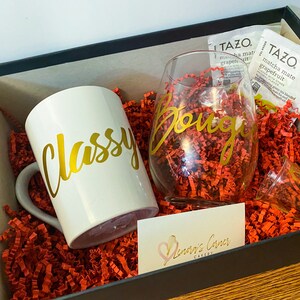 May include: A gift box with a white mug that says "Classy" in gold script, a wine glass with "Bougie" in gold script, and a small shot glass. The box also contains two tea packets and red paper filler.