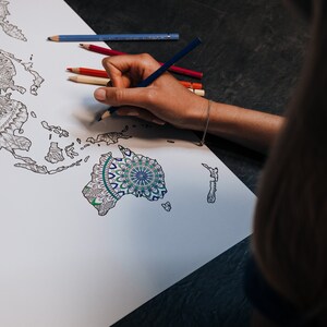 May include: A person coloring in a world map with a blue colored pencil. The map is printed with a mandala design in the center of Australia.