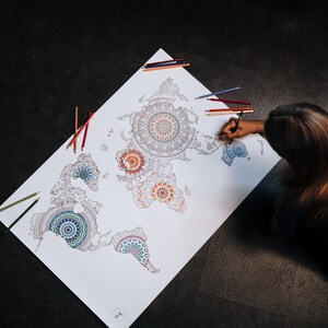 May include: A white world map with intricate mandala designs in various colors, including blue, green, red, and orange. A person is coloring the map with colored pencils.