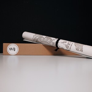 May include: A rolled up white paper map with a black and white mandala design. The map is tied with a black ribbon and is sitting on a brown cardboard box with the text "Mandala Maps Since 2020" and a stylized "M" logo.
