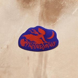 May include: A square-shaped drum with a light beige, textured skin surface. Centered on the drum is a blue sticker featuring a red graphic of a figure flying on a broomstick, a crescent moon, and the text REVULÇAO in a stylized red font.