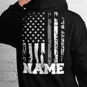 May include: Black hooded sweatshirt with a white distressed American flag design on the back. The flag has a thin gray stripe down the middle and the word "NAME" is printed below the flag.