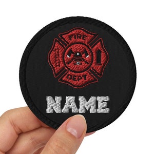 May include: Black circular patch featuring a red embroidered firefighter's Maltese cross design and the text "FIRE DEPT 1". The word "NAME" is embroidered in white below the design. This patch is likely for a firefighter's uniform.