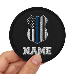 Puede incluir: Parche bordado negro con una bandera americana de línea azul y un escudo blanco. El texto "NAME" está bordado debajo de la bandera.