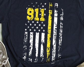 May include: Navy blue t-shirt featuring a distressed American flag design with a yellow stripe and the number "911" in yellow. The shirt is paired with gray sneakers and khaki pants, suggesting casual wear.