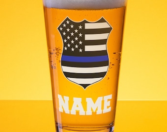 May include: A clear pint glass filled with beer and foam, featuring a shield design with the American flag, a blue line, and the word "NAME" in white. The glass is set against a yellow background.