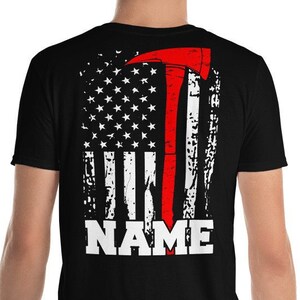 Könnte beinhalten: Schwarzes T-Shirt mit einem abgenutzten amerikanischen Flaggen-Design mit einer roten Feuerwehr-Axt und dem Wort "NAME" in Weiß.