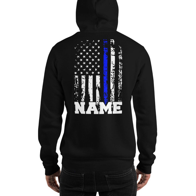 Puede incluir: Sudadera con capucha negra con un dise&ntilde;o de bandera estadounidense desgastado que presenta una l&iacute;nea azul delgada en el centro. El texto "NAME" est&aacute; impreso debajo de la bandera.