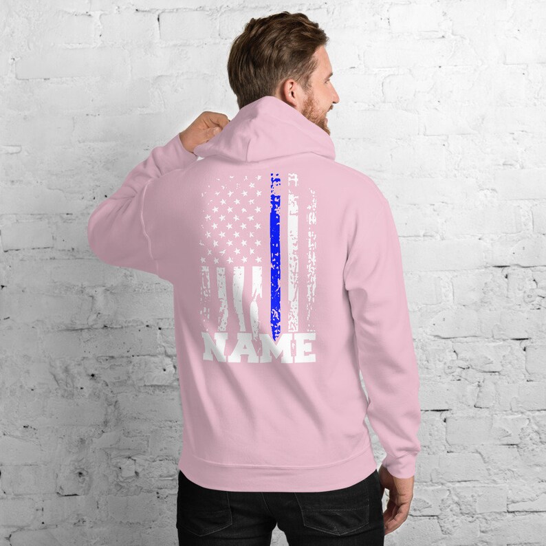 Puede incluir: Sudadera con capucha rosa con un dise&ntilde;o de bandera estadounidense desgastado con una l&iacute;nea azul en el centro. El texto "NAME" est&aacute; impreso en blanco debajo de la bandera.