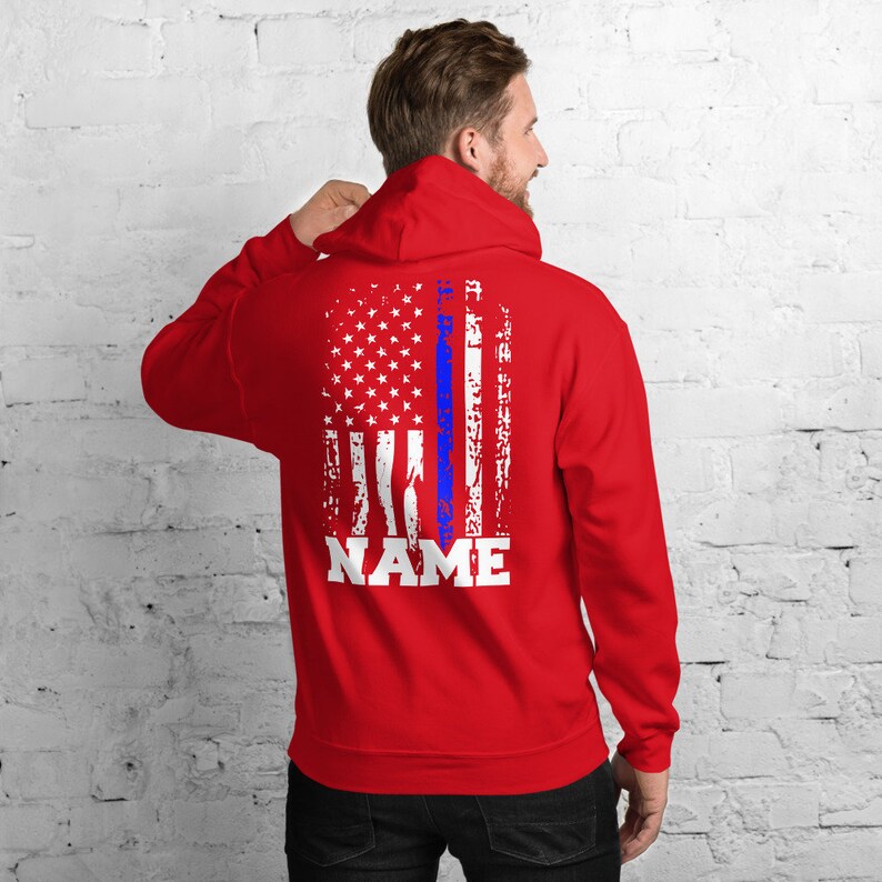 Puede incluir: Sudadera con capucha roja con un dise&ntilde;o de bandera estadounidense desgastado y una fina l&iacute;nea azul en el centro. El texto "NAME" est&aacute; impreso debajo de la bandera.
