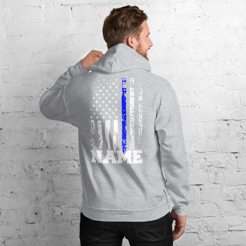 Puede incluir: Una sudadera con capucha gris con un dise&ntilde;o de bandera estadounidense blanca desgastada y una l&iacute;nea azul en el centro. El texto "NAME" est&aacute; impreso debajo de la bandera.