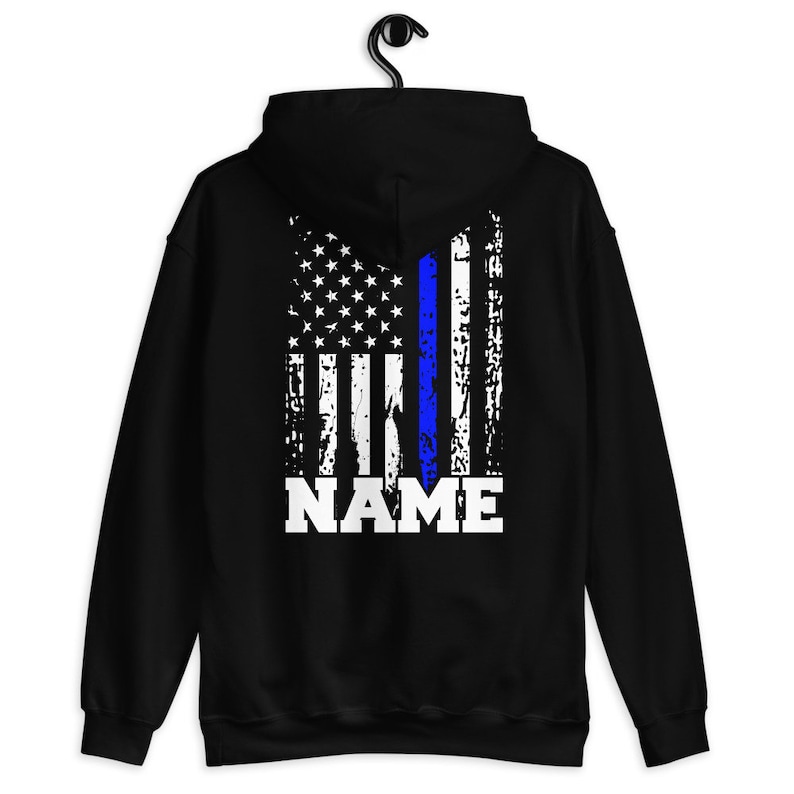 Puede incluir: Sudadera con capucha negra con un dise&ntilde;o de bandera estadounidense desgastado con una l&iacute;nea azul en el centro. El texto "NAME" est&aacute; impreso debajo de la bandera.