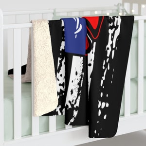 May include: A black, white, red, and blue fleece throw blanket with a distressed graphic design. The blanket is folded and draped over a white crib.