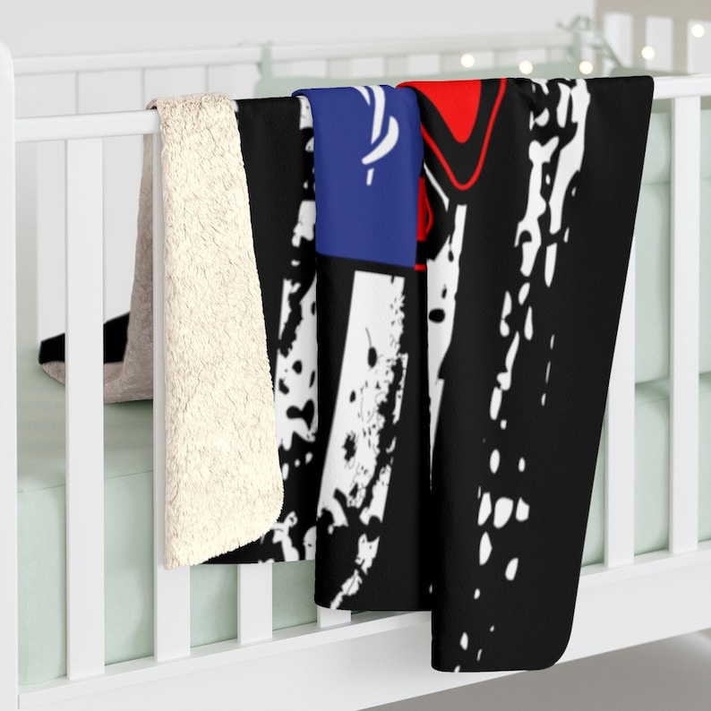 May include: A black, white, red, and blue fleece throw blanket with a distressed graphic design. The blanket is folded and draped over a white crib.
