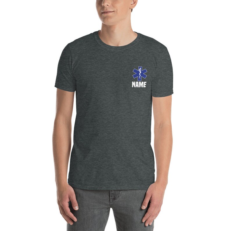 May include: Dark gray t-shirt featuring a blue Star of Life emblem and the word "NAME" in white. The emblem is a six-pointed star with a snake and staff design, commonly associated with emergency medical services.