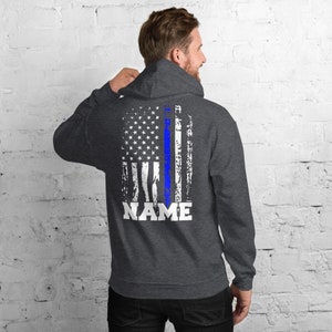 Puede incluir: Una sudadera con capucha gris oscuro con un dise&ntilde;o de bandera estadounidense desgastado que presenta una l&iacute;nea azul en el centro. El texto "NAME" est&aacute; impreso debajo de la bandera.