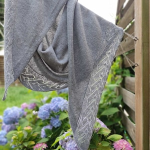 Könnte beinhalten: Ein grauer gestrickter Schal mit einem zarten Spitzenmuster hängt an einem Holzzaun vor einem Hintergrund aus lila und rosa Hortensien.