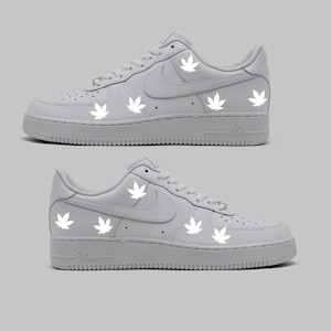 Peut inclure: Baskets basses en cuir blanc avec des motifs de feuilles de marijuana blanches. Les baskets ont un logo Swoosh blanc sur le côté et un logo "AIR" blanc à l'arrière.