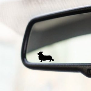Op de afbeelding: Een zwart silhouet van een corgi is zichtbaar in de achteruitkijkspiegel van een auto. De spiegel is zwart en heeft een aan/uit-knop aan de onderkant.