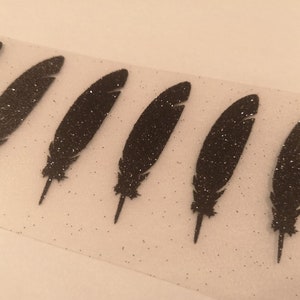 Peut inclure: Une feuille de vinyle thermocollant noir pailleté avec sept formes de plumes découpées. Le vinyle est sur un fond blanc.
