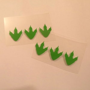 Pode incluir: Cinco pegadas de dinossauro verdes em uma folha de plástico transparente. As pegadas estão dispostas em duas filas, com três pegadas na fila superior e duas pegadas na fila inferior.