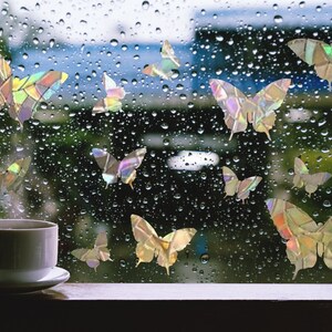 May include: A window with raindrops on it, decorated with iridescent butterfly stickers. A white coffee cup sits on a wooden surface in the foreground.
