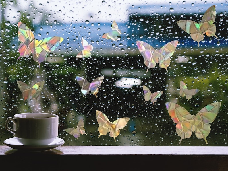 May include: A window with raindrops on it, decorated with iridescent butterfly stickers. A white coffee cup sits on a wooden surface in the foreground.