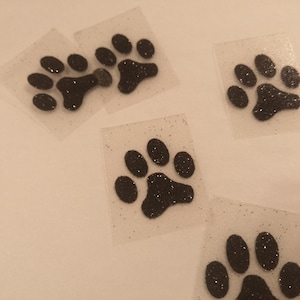 Pode incluir: Cinco patches termocolantes com forma de pata de animal em glitter preto sobre um fundo branco. As pegadas estão em quadrados de plástico transparente.
