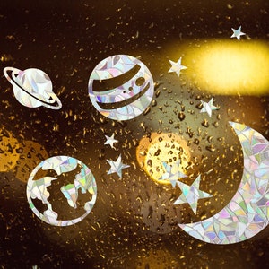 Puede incluir: Una ventana con gotas de lluvia, decorada con pegatinas iridiscentes de planetas, estrellas y una luna creciente.