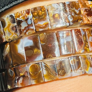 May include: A bracelet made of brown, white and gold agate stones. The stones are cut into square shapes and are strung together on a clear elastic cord.
