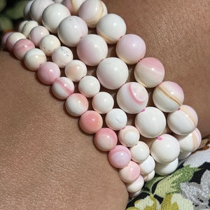 Peut inclure: Cinq bracelets superposés en perles de coquillage rose et blanc. Les perles sont rondes et ont une finition lisse et polie.