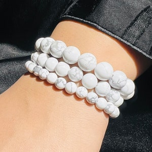Könnte beinhalten: Drei weiße Howlith-Perlenarmbänder, die auf dem Handgelenk einer Person übereinander liegen. Die Perlen sind rund und haben ein marmoriertes Muster.