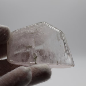 Könnte beinhalten: Ein klarer, durchscheinender, blassrosa Kristall mit geometrischer Form. Der Kristall hat eine glatte, polierte Oberfläche und scheint ein natürliches Mineral zu sein.