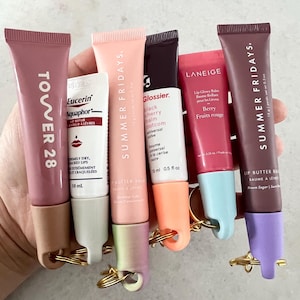 May include: A collection of seven lip balms in various colours and brands, including Tower 28, Summer Fridays, Glossier, Laneige, and Eucerin. The lip balms are attached to keychains.