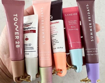 May include: A collection of seven lip balms in various colors and brands, including Tower 28, Summer Fridays, Glossier, Laneige, and Eucerin. The lip balms are attached to keychains.