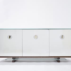 Puede incluir: Un aparador de tres puertas con espejo, tapa de cristal y herrajes de latón. El aparador está hecho de madera con un acabado marrón oscuro.